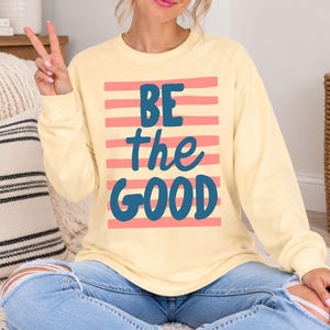 May include: A butter yellow long-sleeve crewneck sweatshirt featuring a graphic print of horizontal salmon pink stripes and the dark blue text "BE the GOOD" centered on the front. The garment has a ribbed neckline, cuffs, and hem.