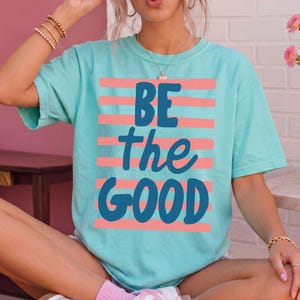 May include: A light turquoise short-sleeve t-shirt featuring horizontal coral pink stripes and the dark blue text "BE the GOOD" printed on the front. The shirt has a crew neckline and a relaxed fit, showcasing a casual and comfortable design.