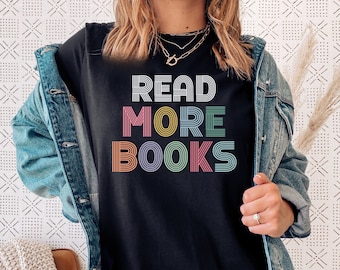 May include: A black short-sleeved t-shirt featuring the text READ MORE BOOKS in a multi-colored, striped, retro-inspired font. The shirt is worn under a blue denim jacket. The graphic text is centered on the chest in shades of pink, yellow, and teal.