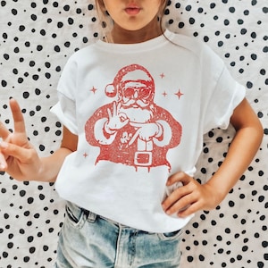 May include: White t-shirt with a red graphic of Santa Claus wearing sunglasses and making a peace sign. The shirt has a crew neck and short sleeves. The word "WHITE" is in a white circle in the upper left corner.