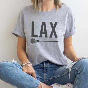 May include: A gray t-shirt with the word "LAX" printed in black on the front. A black lacrosse stick is printed below the word. The shirt is being worn by a person sitting down with their legs crossed.