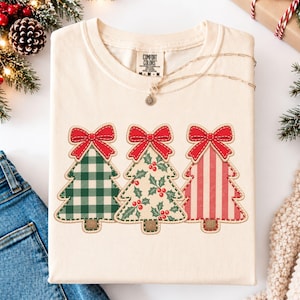 May include: An ivory t-shirt featuring a graphic of three stylized Christmas trees, each topped with a red bow. The first tree has a green and white gingham pattern, the second a holly leaf and berry print, and the third has red and white stripes.