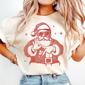 May include: Ivory-colored t-shirt featuring a red graphic of Santa Claus wearing sunglasses and making an "okay" hand gesture. The Santa graphic is surrounded by star shapes. The shirt has short sleeves and a crew neck.