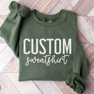 May include: A military green sweatshirt with the words "CUSTOM" above "sweatshirt" in white. The sweatshirt has a crew neck and long sleeves with ribbed cuffs. The words "MILITARY GREEN" are in a circle at the bottom left.
