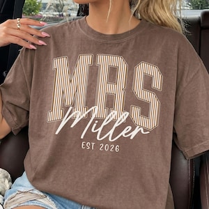 May include: A brown short-sleeve t-shirt featuring the text MRS in large, beige-striped block letters. Below this, the word Miller is printed in a white script font, followed by the text EST 2026 in a simple, clean white sans-serif typeface.