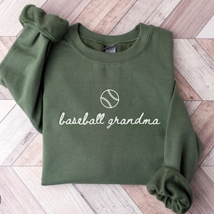 May include: A military green sweatshirt with the words "baseball grandma" in white script, and a white baseball graphic. The sweatshirt has a crew neck and long sleeves with ribbed cuffs. The words "MILITARY GREEN" are in a circle at the bottom left.