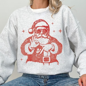 May include: A light gray sweatshirt featuring a red graphic of Santa Claus wearing sunglasses and making an 'okay' hand gesture. The Santa graphic has a distressed, vintage look. The sweatshirt is a casual, long-sleeved top.