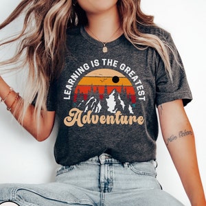 May include: Dark grey heather t-shirt with a vintage-style graphic that reads "Learning is the greatest adventure". The graphic features a sunset with mountains and trees.
