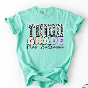 May include: A light turquoise t-shirt with the words "THIRD GRADE Mrs. Anderson" printed on the front. The word "THIRD" is in a black and white pattern, while "GRADE" is in pastel colors. The shirt is a comfortable, casual style.