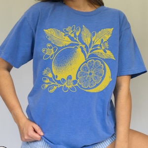 Op de afbeelding: Een blauw T-shirt met een gele citroondruk. De citroen is omringd door bladeren en bloemen.