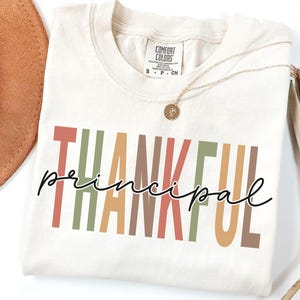 May include: Ivory-colored t-shirt with the words "THANKFUL principal" in a mix of colors. The word "THANKFUL" is in a bold, block letter style, while "principal" is in a cursive font. A gold necklace with a pendant is draped across the shirt.