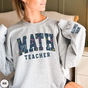 May include: A gray sweatshirt with the text "MATH TEACHER" in navy blue lettering. The word "MATH" is filled with colorful math symbols. The sleeve has the text "MRS. SMITH" in navy blue lettering.