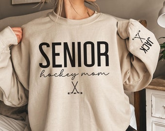 May include: A beige sweatshirt with the text "SENIOR hockey mom" and a pair of crossed hockey sticks. The right sleeve has the text "JACK" and a pair of crossed hockey sticks.