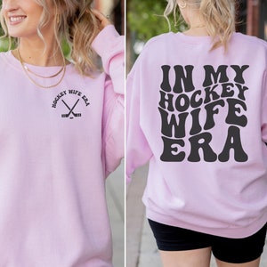 Pu&ograve; includere: Una felpa rosa chiaro con il testo "In My Hockey Wife Era" in lettere nere in stile retr&ograve; sul retro. La parte anteriore della felpa presenta una piccola grafica di bastoni da hockey incrociati con il testo "Hockey Wife Era" in un cerchio.