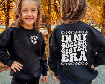 May include: Black sweatshirt with white text that says "In My Soccer Sister Era" and a soccer ball graphic. The sweatshirt also has a white soccer ball graphic and the text "Miller" on the front.
