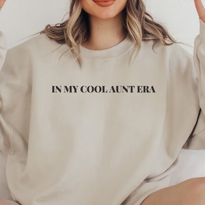 May include: A beige sweatshirt with the text "IN MY COOL AUNT ERA" in black block letters. The sweatshirt has long sleeves and a crew neck. The overall style is casual and comfortable.