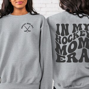 May include: A gray sweatshirt with the text "In My Hockey Mom Era" in a retro font on the back. The front of the sweatshirt has a small graphic of crossed hockey sticks with the text "Hockey Mom Era" around the sticks.