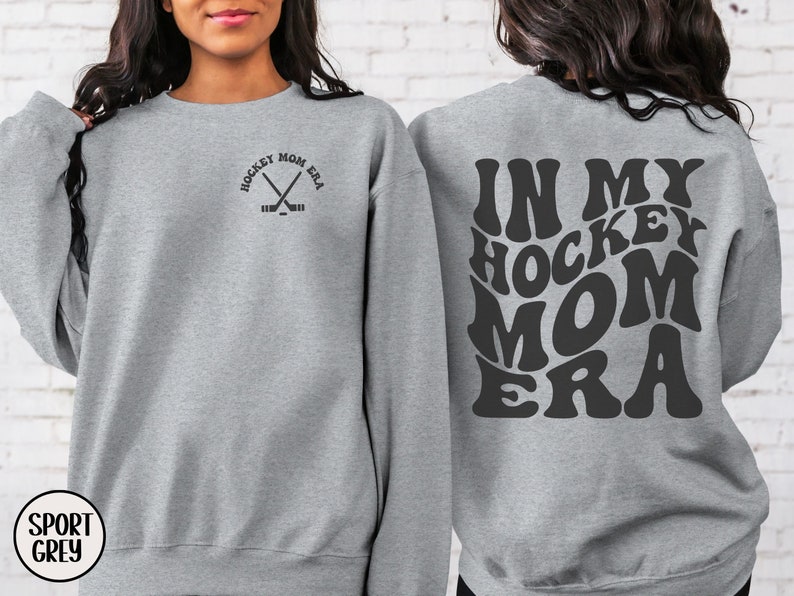 May include: A gray sweatshirt with the text "In My Hockey Mom Era" in a retro font on the back. The front of the sweatshirt has a small graphic of crossed hockey sticks with the text "Hockey Mom Era" around the sticks.
