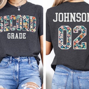 Puede incluir: Camiseta gris oscuro con la inscripci&oacute;n "SECOND GRADE" en un dise&ntilde;o floral en la parte delantera. La parte trasera de la camiseta muestra "JOHNSON 02" con detalles florales. La camiseta se combina con vaqueros azules.