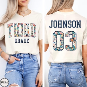 May include: A cream-colored t-shirt featuring the text THIRD GRADE on the front in dark blue block letters with a colorful floral pattern inside. The back displays the name JOHNSON and the number 03 in the same dark blue and floral style.