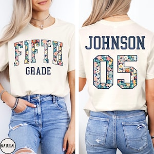 Op de afbeelding: Cr&egrave;mekleurig T-shirt met "FIFTH GRADE" in een bloemenontwerp op de voorkant. De achterkant heeft "JOHNSON 05" in een vergelijkbaar bloemenpatroon. Het shirt wordt gedragen met een spijkerbroek.