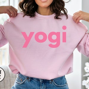 Może przedstawiać: R&oacute;żowa bluza z nadrukiem słowa "yogi" w dużym, pogrubionym, r&oacute;żowym foncie.