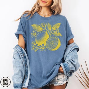 Op de afbeelding: Een jeansblauw T-shirt met een gele citroenprint. De citroen is omringd door bladeren en bloemen.