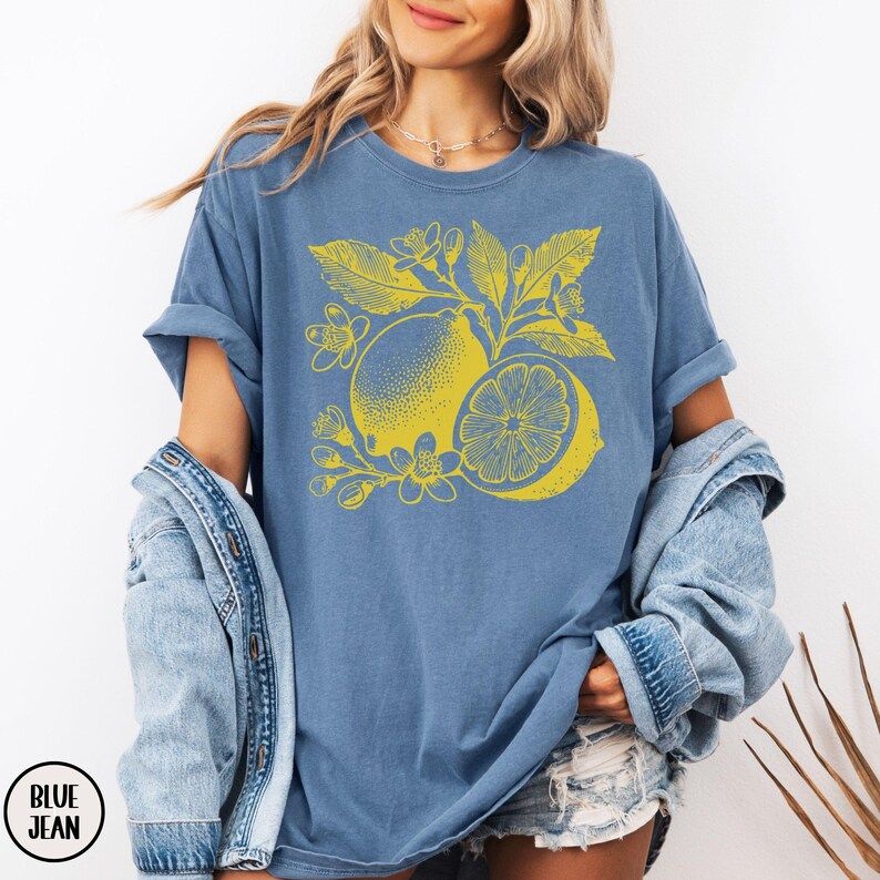 Op de afbeelding: Een jeansblauw T-shirt met een gele citroenprint. De citroen is omringd door bladeren en bloemen.