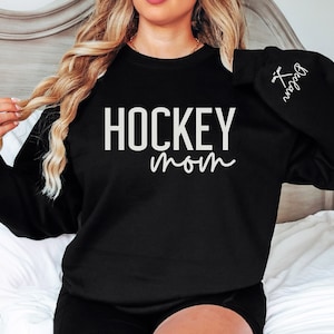 May include: Black sweatshirt with the text "HOCKEY mom" in white lettering. The right sleeve has a small white hockey stick graphic with the text "Hockey Mom" in white lettering.