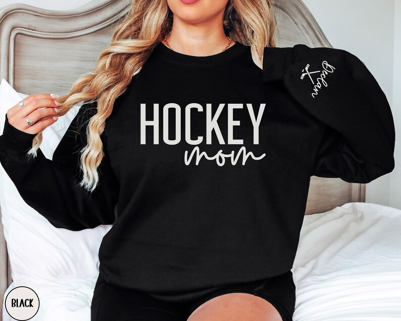 May include: Black sweatshirt with the text "HOCKEY mom" in white lettering. The right sleeve has a small white hockey stick graphic with the text "Hockey Mom" in white lettering.