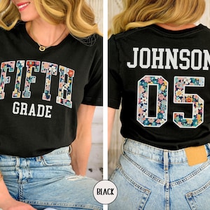 Op de afbeelding: Zwart T-shirt met de woorden "FIFTH GRADE" in een bloemenpatroon op de voorkant. De achterkant van het shirt toont "JOHNSON 05" met een bloemenpatroon in het nummer. Het shirt wordt gedragen met een blauwe jeans.