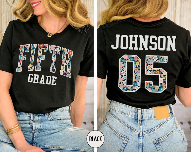 Op de afbeelding: Zwart T-shirt met de woorden "FIFTH GRADE" in een bloemenpatroon op de voorkant. De achterkant van het shirt toont "JOHNSON 05" met een bloemenpatroon in het nummer. Het shirt wordt gedragen met een blauwe jeans.