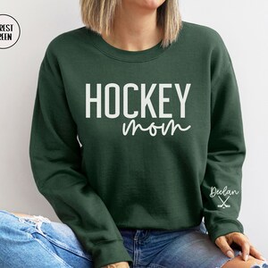 May include: Forest green sweatshirt with the text "HOCKEY mom" in white lettering. The right sleeve has the name "Declan" and a pair of hockey sticks in white.