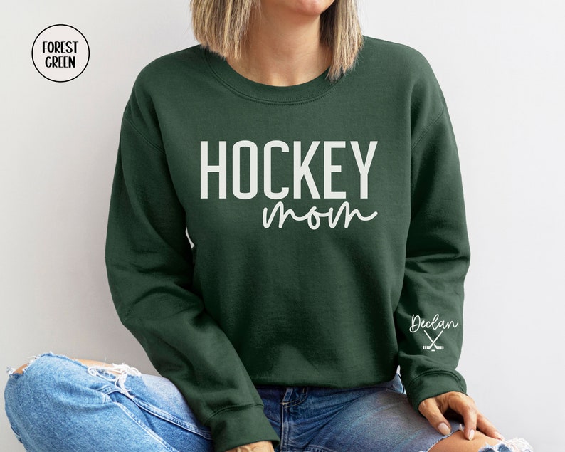 May include: Forest green sweatshirt with the text "HOCKEY mom" in white lettering. The right sleeve has the name "Declan" and a pair of hockey sticks in white.