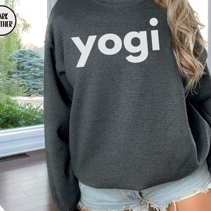 Może przedstawiać: Szara bluza z kapturem w kolorze ciemnego heathered z napisem "yogi" nadrukowanym na przodzie w kolorze białym.