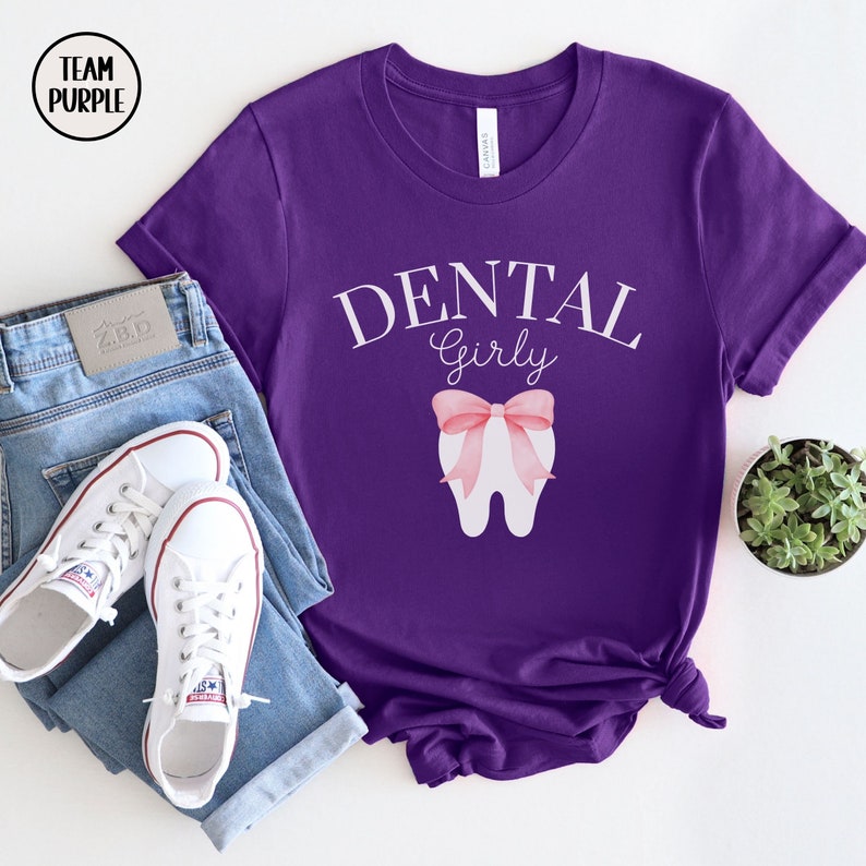 May include: Purple t-shirt with the text "Dental Girly" in white lettering. A white tooth with a pink bow is centered on the shirt.