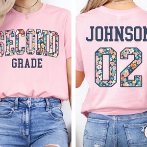 Puede incluir: Camiseta rosa claro con las palabras "SECOND GRADE" en un dise&ntilde;o floral en la parte delantera. La parte trasera muestra el nombre "JOHNSON" y el n&uacute;mero "02", tambi&eacute;n en dise&ntilde;o floral.