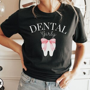 May include: A black t-shirt with the text "Dental Girly" in white lettering. A pink tooth with a pink bow is centered on the shirt.