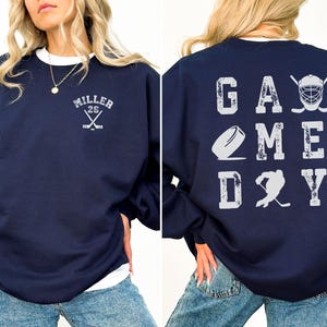 May include: Navy blue sweatshirt with a white graphic on the back that says "GAME DAY" with hockey-themed imagery. The front of the sweatshirt has a white graphic with the text "MILLER 26" and crossed hockey sticks.