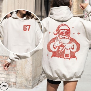 May include: Sand-colored hoodie with a red graphic of Santa Claus making an 'okay' hand gesture. The front of the hoodie features the number '67' in red. The hoodie has a drawstring hood and a kangaroo pocket.