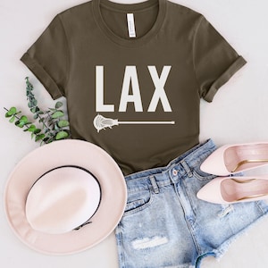 May include: A green t-shirt with white text that says "LAX" and a white lacrosse stick graphic. The shirt is paired with denim shorts and a pink hat.