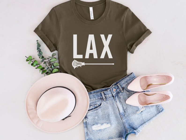 May include: A green t-shirt with white text that says "LAX" and a white lacrosse stick graphic. The shirt is paired with denim shorts and a pink hat.