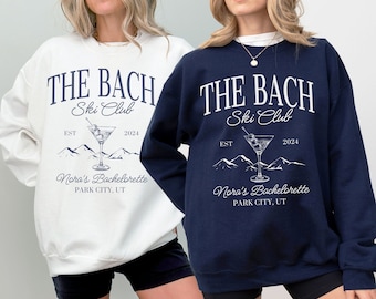 May include: Two white and navy blue sweatshirts with the text "THE BACH Ski Club" and "EST 2024" printed on the front. The design also includes a martini glass with olives, mountains, and the text "Nora's Bachelorette PARK CITY, UT".