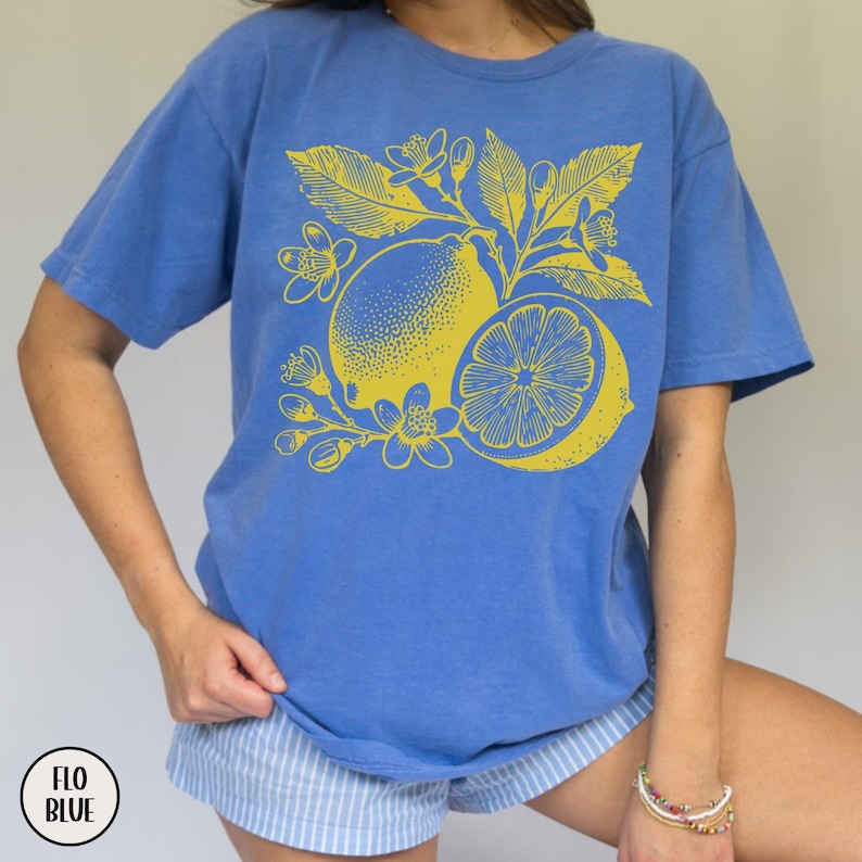 Op de afbeelding: Een blauw T-shirt met een gele citroondruk. De citroen is omringd door bladeren en bloemen.