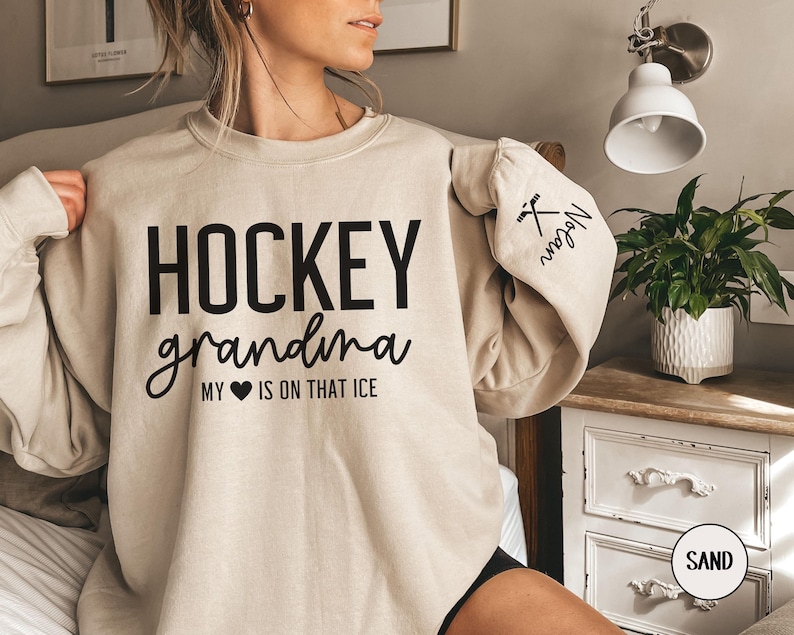 May include: A beige sweatshirt with the text "HOCKEY grandma MY HEART IS ON THAT ICE" printed in black. The left sleeve has a black hockey stick and puck graphic with the text "Megan" printed in black.