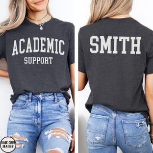 May include: Dark grey heather t-shirt with the words "ACADEMIC SUPPORT" printed in white on the front. The back of the shirt displays the name "SMITH" in white block letters. The model is wearing blue jeans.