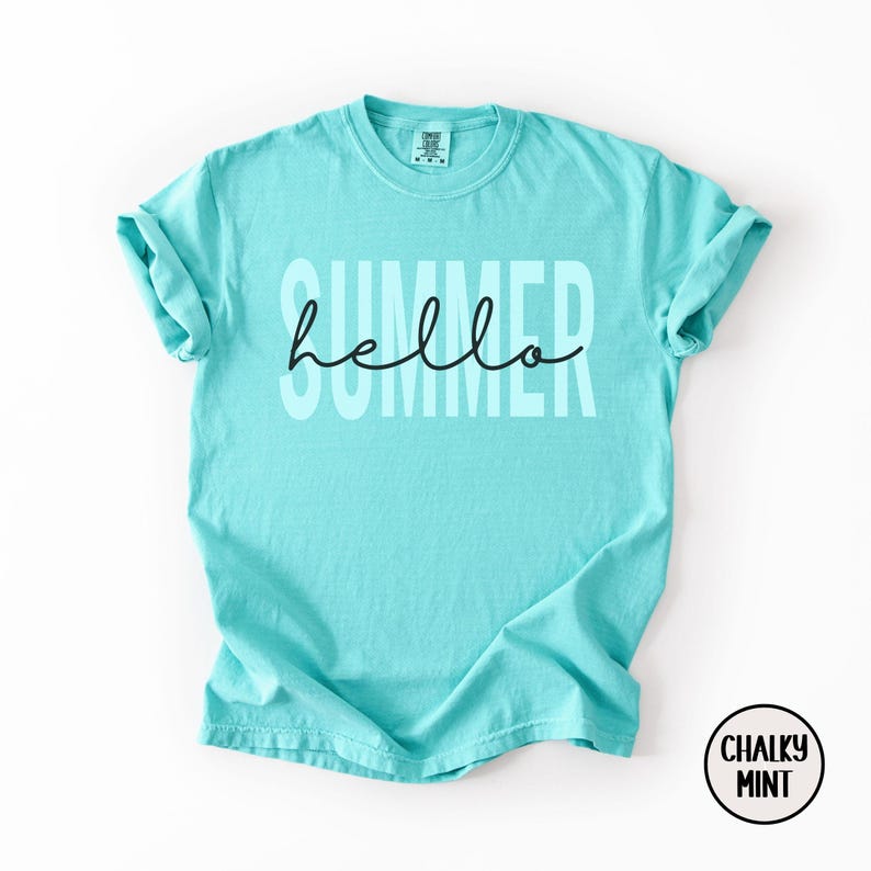 May include: A turquoise t-shirt with the word "SUMMER" in large, light-colored block letters and the word "hello" in a black cursive font. The shirt has short sleeves and a crew neck. A circular logo with the words "CHALKY MINT" is in the bottom right corner.