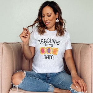Peut inclure: Une femme portant un t-shirt gris avec le texte "TEACHING is my JAM" et un graphique de trois tranches de pain dessin&eacute;es avec de la confiture et du beurre de cacahu&egrave;te. Elle est assise sur un canap&eacute; rose.