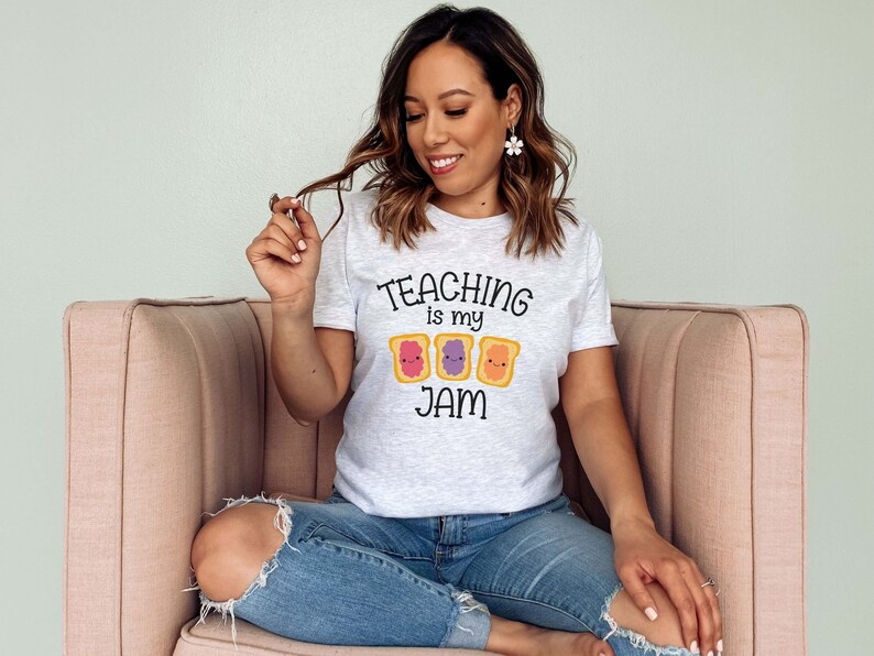 Peut inclure: Une femme portant un t-shirt gris avec le texte "TEACHING is my JAM" et un graphique de trois tranches de pain dessin&eacute;es avec de la confiture et du beurre de cacahu&egrave;te. Elle est assise sur un canap&eacute; rose.
