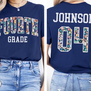 May include: Navy blue t-shirt with "FOURTH GRADE" in white block letters. The word "FOURTH" is filled with a floral pattern. The back of the shirt has "JOHNSON 04" in white block letters, with the "04" also filled with the floral pattern.